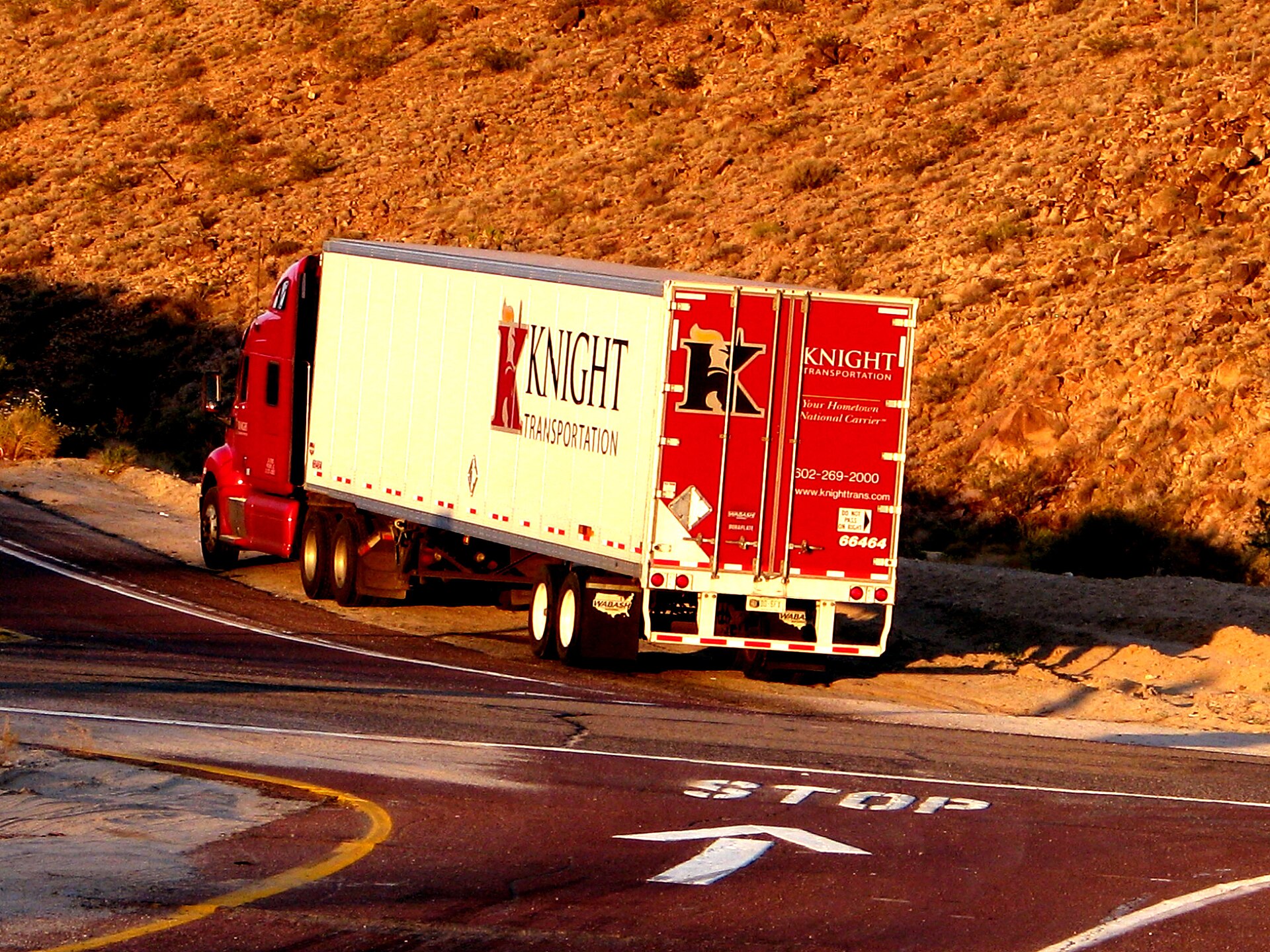 A red semi-truck with a loaded trailer on a two-lane road through open country